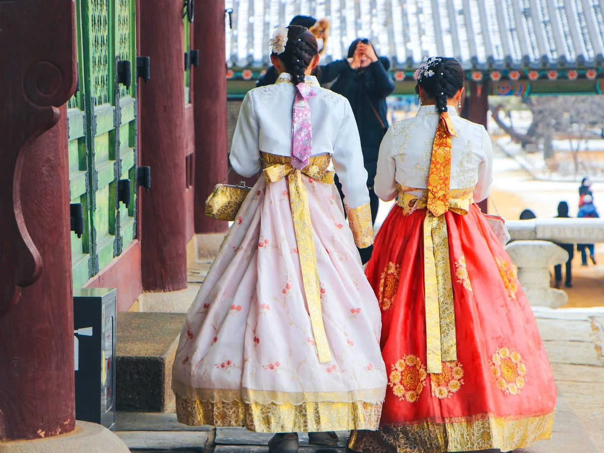 Zwei Personen in traditionellen koreanischen Hanbok-Kleidern stehen vor einem historischen Geb&auml;ude, eine in einem pastellfarbenen Kleid mit gelber Sch&auml;rpe und eine in einem roten Kleid mit goldener Verzierung, vor einer traditionellen koreanischen Architektur.