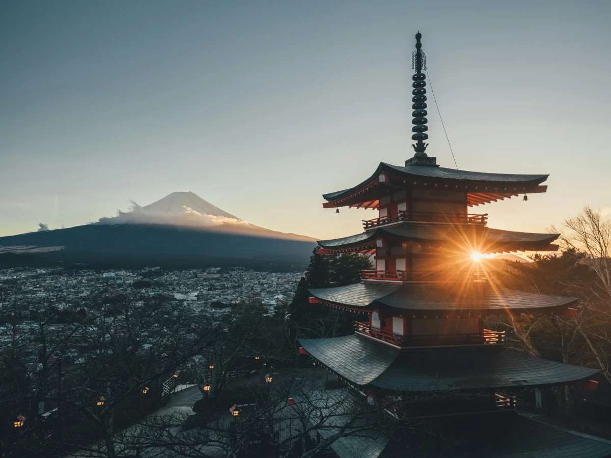 Chureito-Pagode mit Blick auf den schneebedeckten Fuji-Berg bei Sonnenaufgang. Mehrere Stockwerke des roten Tempels im Vordergrund, mit Sonnenstern, der zwischen den Dachebenen hindurchscheint. Weite Stadtlandschaft und B&auml;ume im Hintergrund unter einem klaren Morgenhimmel.