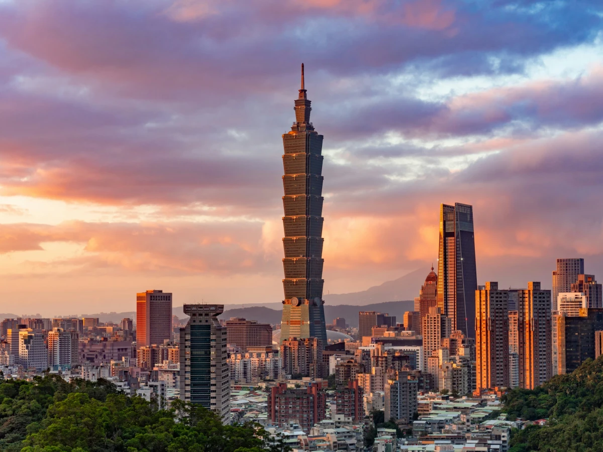 Skyline von Taipeh bei Sonnenuntergang mit dem markanten Taipei 101 Wolkenkratzer im Vordergrund, umgeben von modernen Hochh&auml;usern und einem dramatischen Himmel in Pastellfarben, im Hintergrund bewaldete Berge.