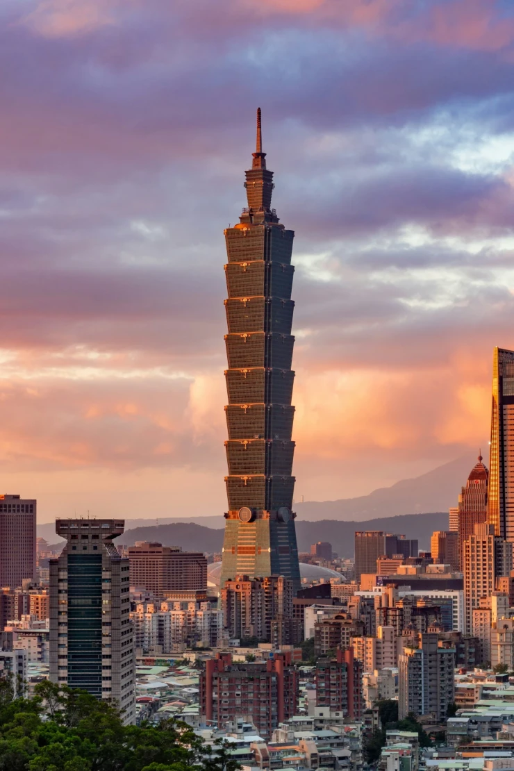 Skyline von Taipeh bei Sonnenuntergang mit dem markanten Taipei 101 Wolkenkratzer im Vordergrund, umgeben von modernen Hochh&auml;usern und einem dramatischen Himmel in Pastellfarben, im Hintergrund bewaldete Berge.
