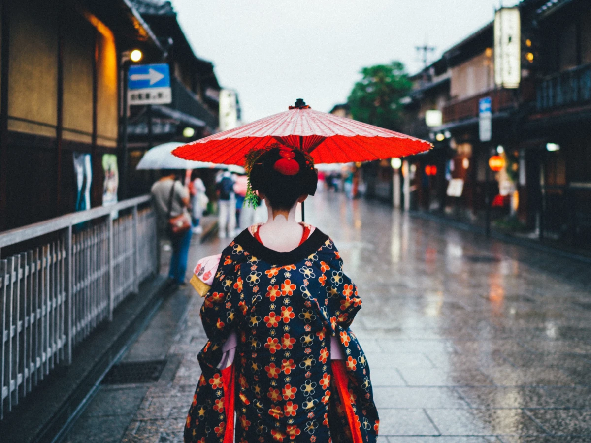 R&uuml;ckansicht einer Person in traditioneller japanischer Kleidung mit einem roten Schirm, die an einem regnerischen Tag eine alte Stra&szlig;e in Kyoto entlang geht, umgeben von historischen Holzgeb&auml;uden und ged&auml;mpften Lichtern.