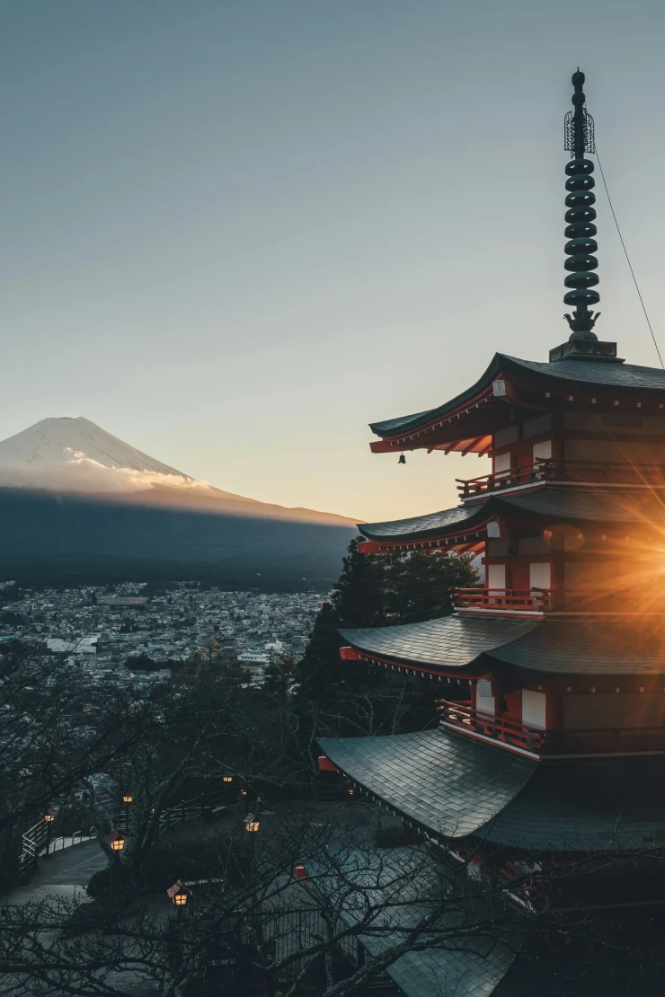 Chureito-Pagode mit Blick auf den schneebedeckten Fuji-Berg bei Sonnenaufgang. Mehrere Stockwerke des roten Tempels im Vordergrund, mit Sonnenstern, der zwischen den Dachebenen hindurchscheint. Weite Stadtlandschaft und B&auml;ume im Hintergrund unter einem klaren Morgenhimmel.