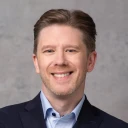 L&auml;chelnder Gesch&auml;ftsmann mittleren Alters im dunklen Blazer und hellblauem Hemd vor einem grauen Hintergrund. Er hat braunes, zur&uuml;ckgek&auml;mmtes Haar und einen freundlichen, selbstbewussten Gesichtsausdruck.