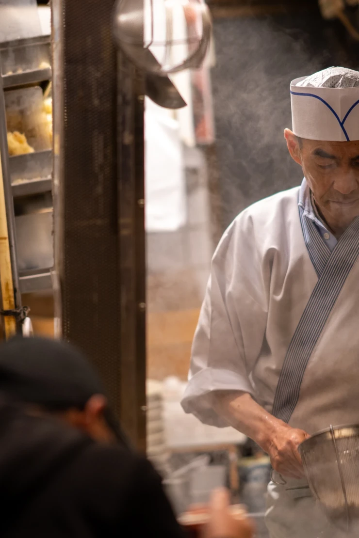 Wei&szlig;gekleideter Koch in einer traditionellen japanischen Ramenk&uuml;che bereitet sorgf&auml;ltig Nudeln zu, umgeben von K&uuml;chenger&auml;ten und ged&auml;mpfter Beleuchtung. Die Atmosph&auml;re ist gesch&auml;ftig und authentisch, mit Blick auf die Zubereitung der Speisen.