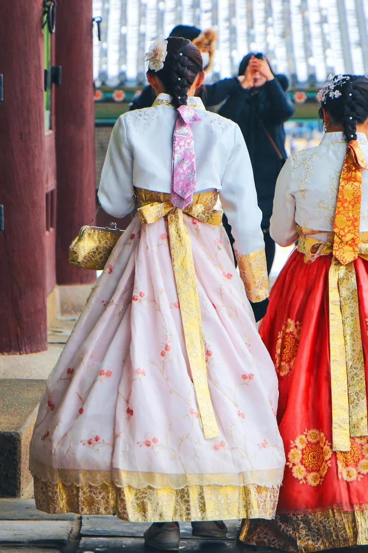 Zwei Personen in traditionellen koreanischen Hanbok-Kleidern stehen vor einem historischen Geb&auml;ude, eine in einem pastellfarbenen Kleid mit gelber Sch&auml;rpe und eine in einem roten Kleid mit goldener Verzierung, vor einer traditionellen koreanischen Architektur.