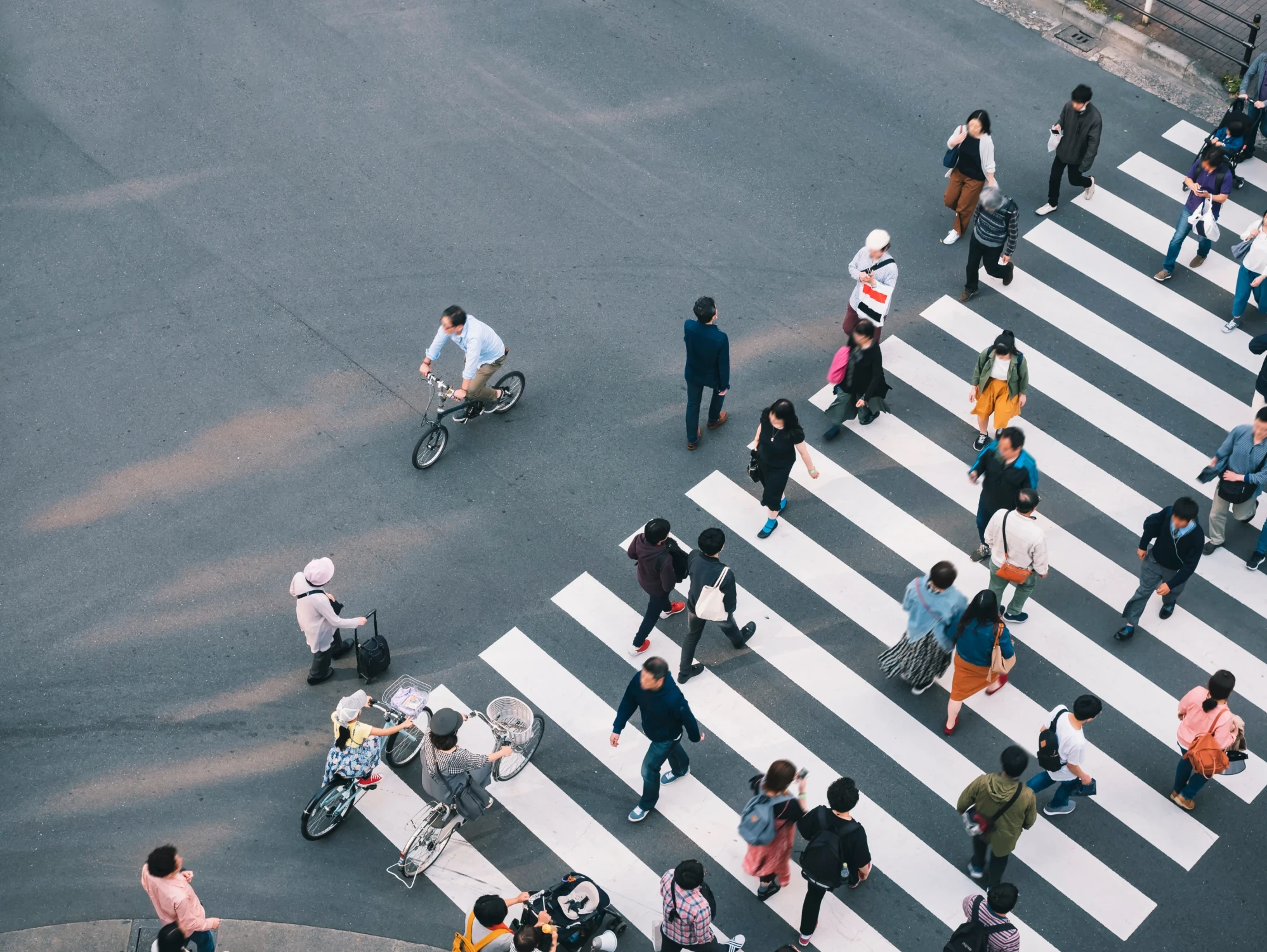 Luftaufnahme einer belebten Fu&szlig;g&auml;nger&uuml;berquerung in einer Stadt mit Menschen in verschiedenen Kleidungsfarben, die einen Zebrastreifen &uuml;berqueren, w&auml;hrend ein Radfahrer im Hintergrund f&auml;hrt.