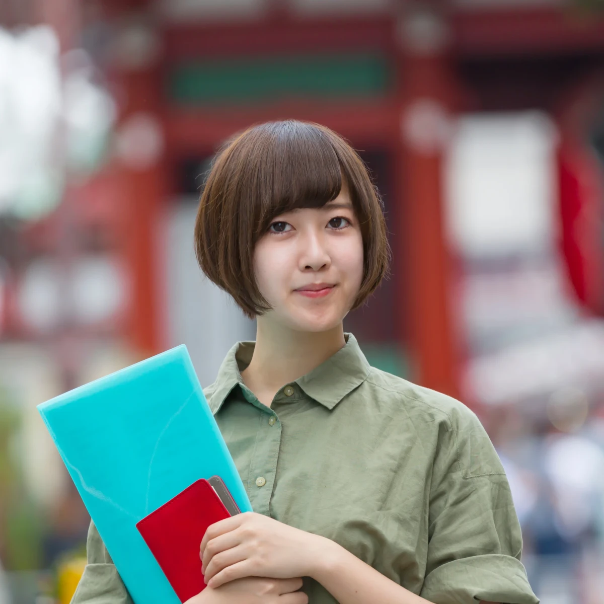 Junge Frau mit Bob-Frisur in olivgr&uuml;nem Hemd steht vor unscharfem rotem Hintergrund und h&auml;lt ein t&uuml;rkis-rotes Buch. Sie blickt freundlich und selbstbewusst in die Kamera, im Hintergrund ist eine verschwommene st&auml;dtische Umgebung zu erkennen.