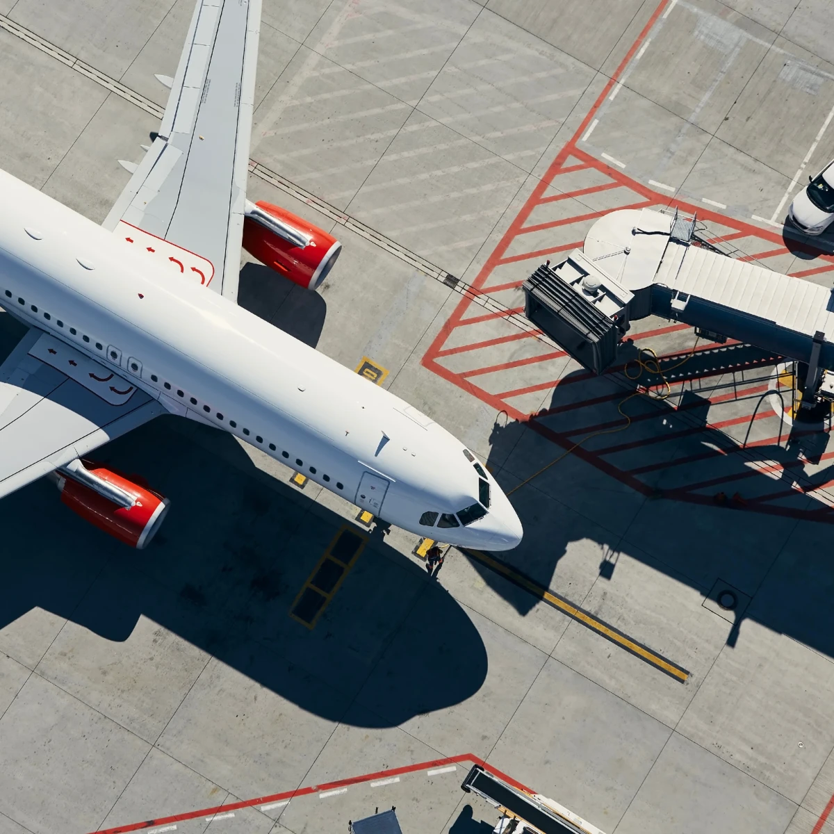 Luftaufnahme eines wei&szlig;en Passagierflugzeugs an einem Flughafengate mit Flugzeugbr&uuml;cke. Rote Triebwerke und Seitenleitwerk sind sichtbar, w&auml;hrend Bodenpersonal und Fahrzeuge um das Flugzeug herum positioniert sind.