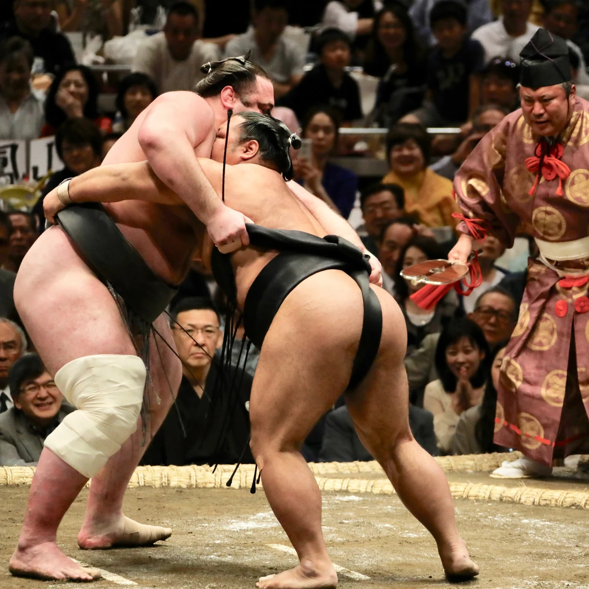 Zwei Sumō-Ringer k&auml;mpfen intensiv auf einem Wettkampfring, umgeben von aufgeregten Zuschauern und einem traditionell gekleideten Schiedsrichter. Der Kampf findet in einer vollen Halle statt, mit Spannung und Konzentration in der Luft.