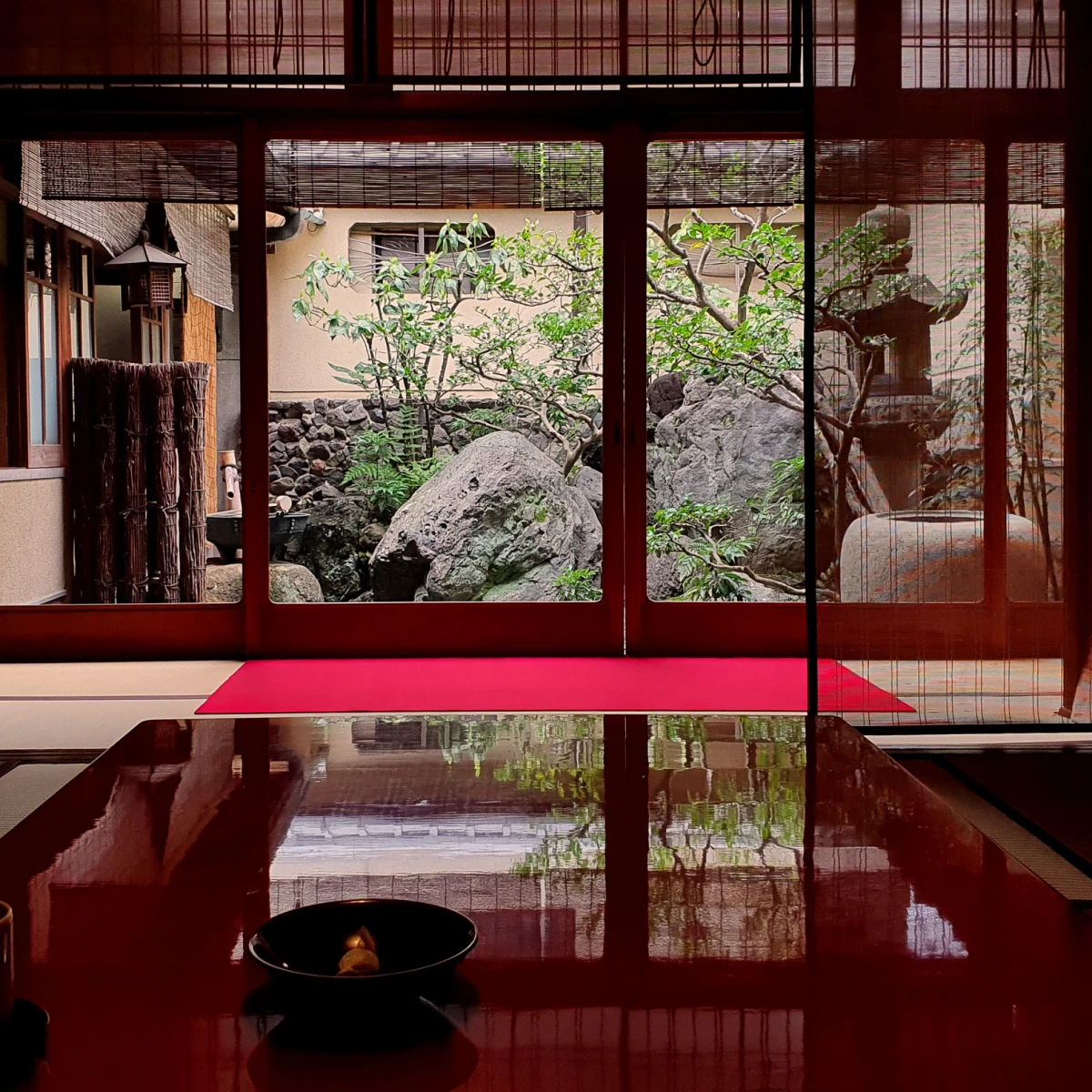 Traditioneller japanischer Raum mit gl&auml;nzend rotem Boden, Schiebet&uuml;ren und Blick auf einen zen-artigen Steingarten mit Baum und Steinlaterne, der Ruhe und Eleganz ausstrahlt.