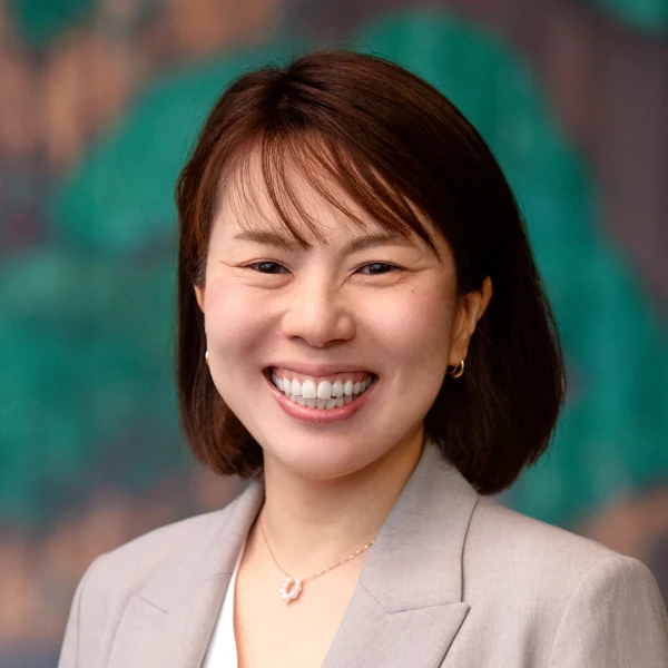 L&auml;chelnde Gesch&auml;ftsfrau in hellem Blazer vor einem gr&uuml;n-braunen unscharfen Hintergrund, mit freundlichem und selbstbewusstem Ausdruck.