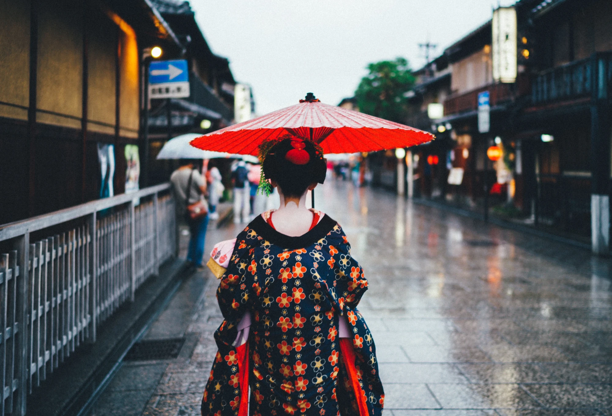 R&uuml;ckansicht einer Person in traditioneller japanischer Kleidung mit einem roten Schirm, die an einem regnerischen Tag eine alte Stra&szlig;e in Kyoto entlang geht, umgeben von historischen Holzgeb&auml;uden und ged&auml;mpften Lichtern.