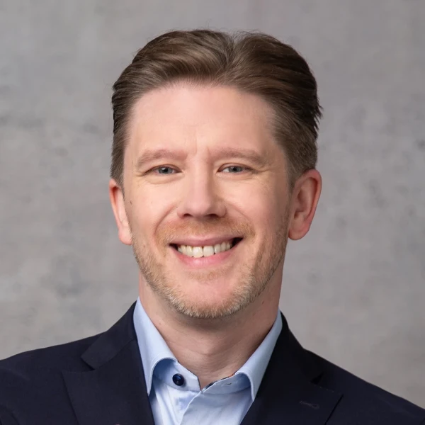 L&auml;chelnder Gesch&auml;ftsmann mittleren Alters im dunklen Blazer und hellblauem Hemd vor einem grauen Hintergrund. Er hat braunes, zur&uuml;ckgek&auml;mmtes Haar und einen freundlichen, selbstbewussten Gesichtsausdruck.