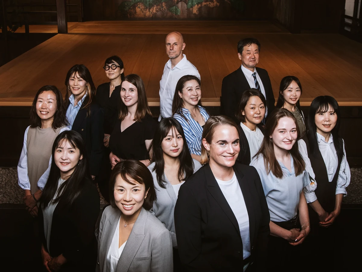 Professionelles Gruppenportr&auml;t eines internationalen Teams mit 13 Mitarbeitenden, das in zwei Reihen posiert und eine Mischung aus M&auml;nnern und Frauen unterschiedlicher Herkunft zeigt, gekleidet in gesch&auml;ftliche Berufskleidung in gedeckten Farbt&ouml;nen vor einem warmen, holzvert&auml;felten Hintergrund.